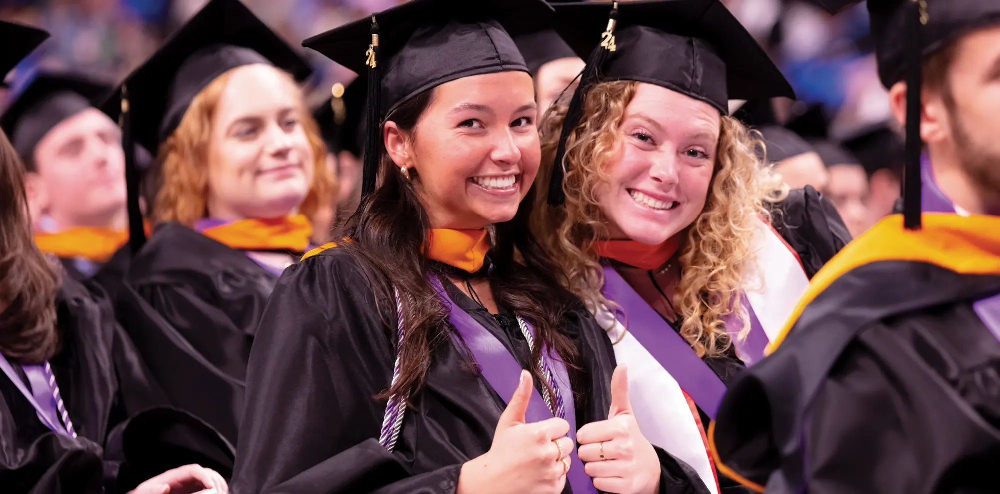 Two smiling graduates in black caps and gowns sit among their classmates during a commencement ceremony