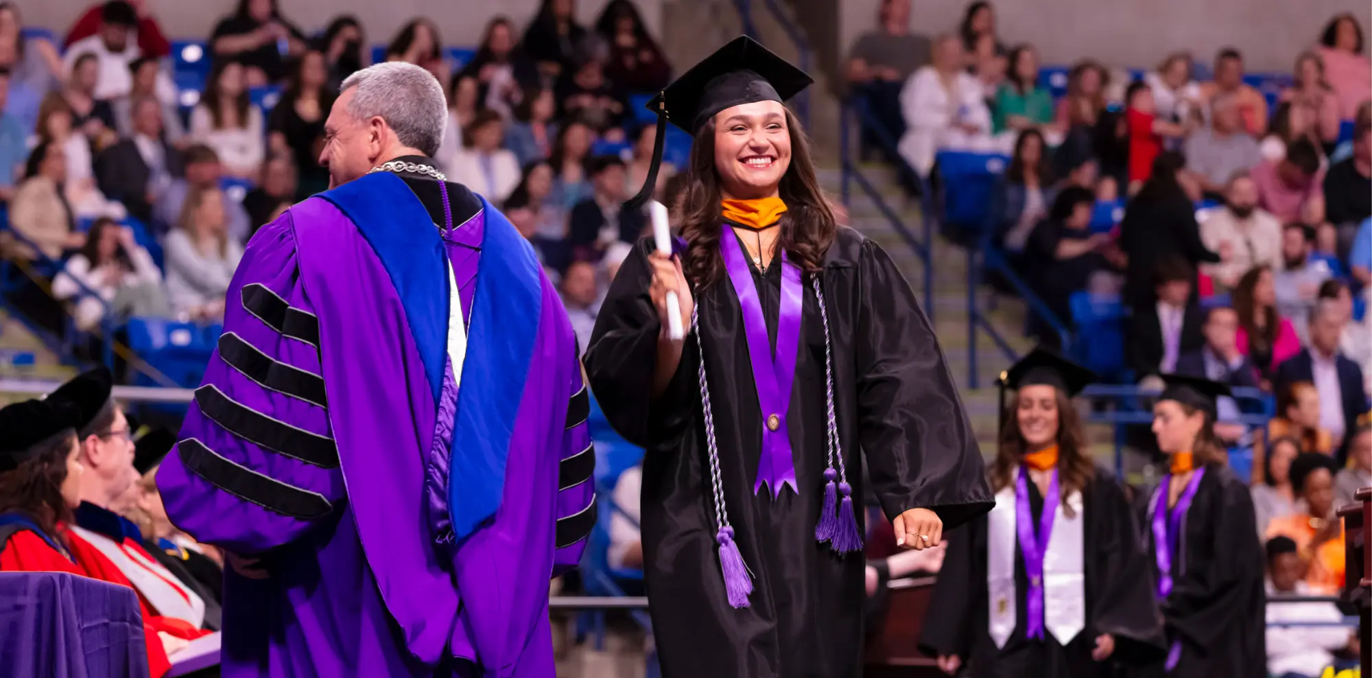 A smiling Scranton graduate in a black gown, cap, and purple honor sashes walks across a stage, holding a diploma, during an indoor graduation ceremony