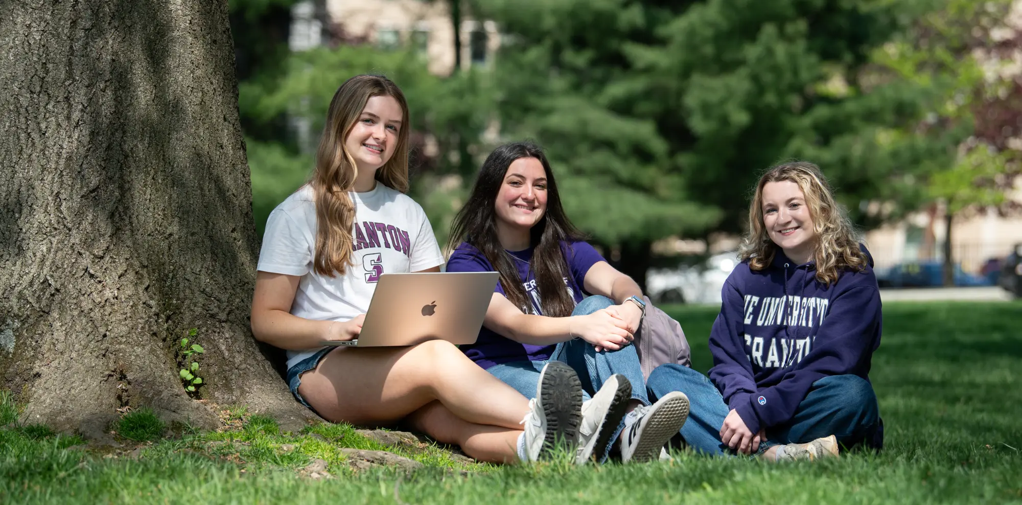 Three smiling Scranton female students sitting on a grassy campus lawn next to a large tree on a sunny day